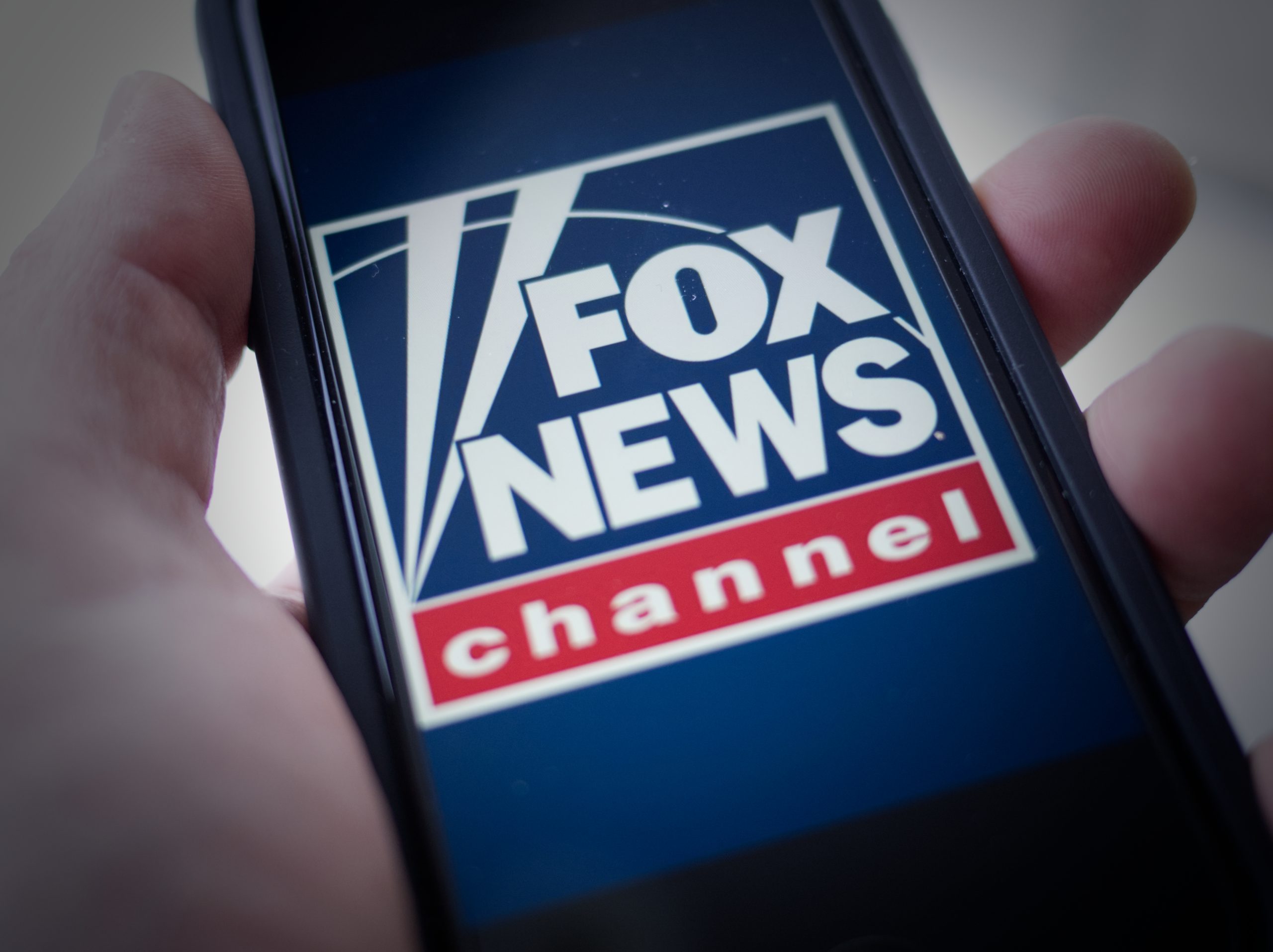 Close-up of a hand holding a mobile phone. On the screen is the logo of Fox News.